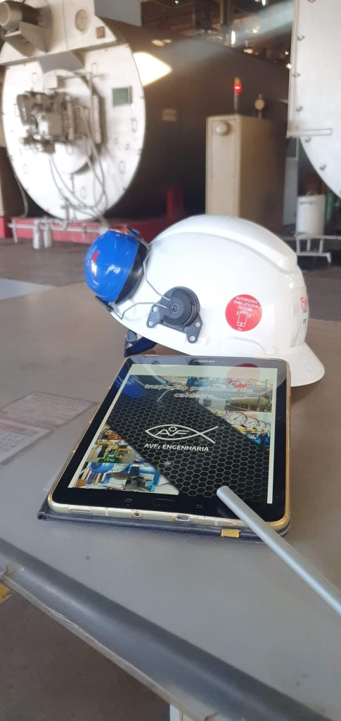 A imagem retrata o contexto de uma inspeção periódica de caldeira em um ambiente industrial. Em destaque, estão um capacete de segurança com protetor auditivo e um tablet que exibe informações relacionadas à inspeção da caldeira, sinalizando a integração de tecnologias digitais no processo de gestão e controle de segurança. O capacete reflete a importância dos Equipamentos de Proteção Individual (EPIs) para garantir a segurança dos trabalhadores em ambientes potencialmente perigosos, como instalações de caldeiras e vasos de pressão.