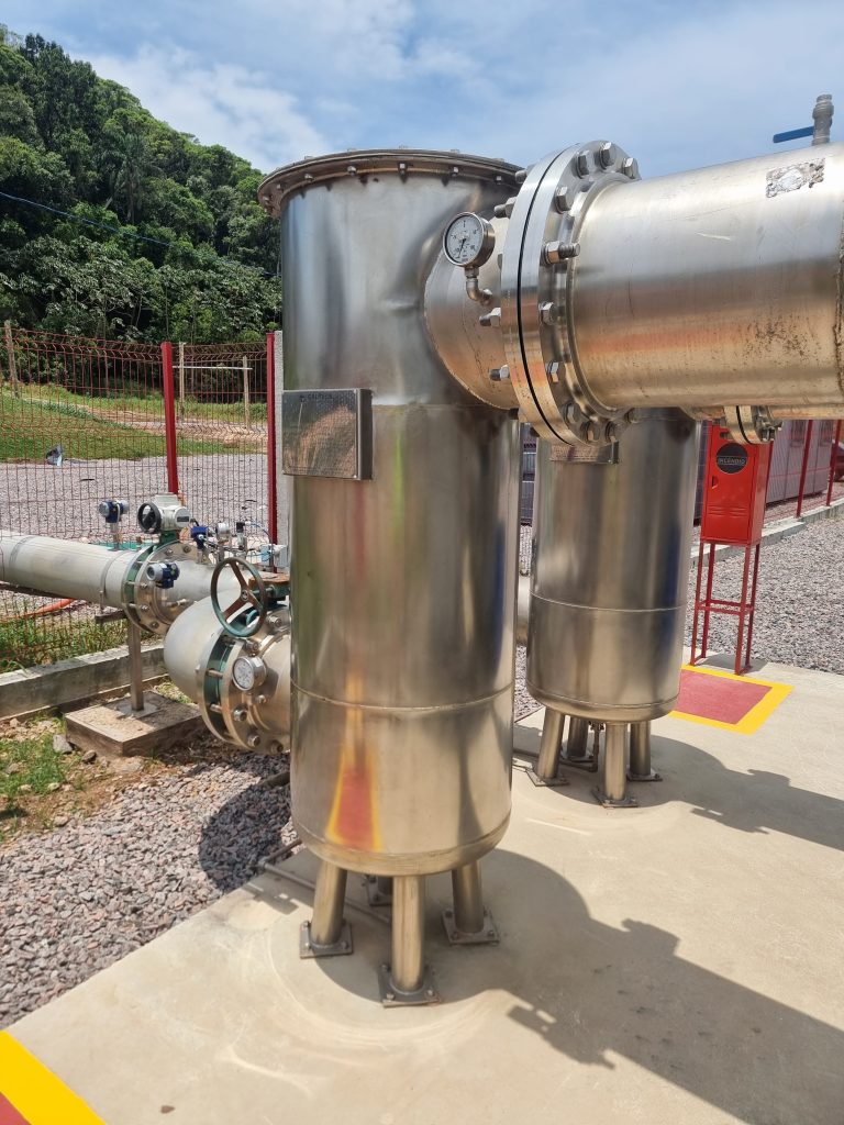 A imagem mostra um vaso de pressão em operação em um ambiente industrial ao ar livre, cercado por uma área de segurança. O equipamento, construído em aço inoxidável, está conectado a tubulações por meio de flanges robustas, e um manômetro é visível, indicando a importância do monitoramento contínuo da pressão interna. Este tipo de vaso é frequentemente utilizado em processos industriais, como nas indústrias química, petroquímica e de energia, para conter substâncias sob pressão, garantindo a segurança e eficiência operacional.